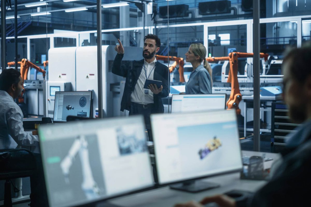 Electric Engine Production At Autonomous Factory With Robotic Arms: Diverse Technicians And Engineers Using Desktop Computers To Control Assembly Line While Managers With Tablet Discussing Project